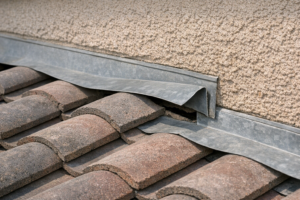 Illustration of a lifted metal roof-to-wall flashing edge beside concrete roof tiles and a stucco wall.